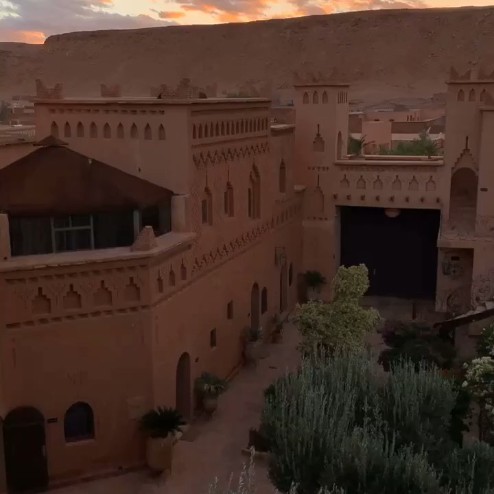 A high-angle view of the historic clay-brick Kasbah Amridil in Skoura, Morocco, during a warm sunset. The traditional earth-toned earthen fortress features crenellated towers, geometric decorative engravings, and arched entryways enclosing a central courtyard garden filled with lush green shrubs and palm trees against a desert hill background.