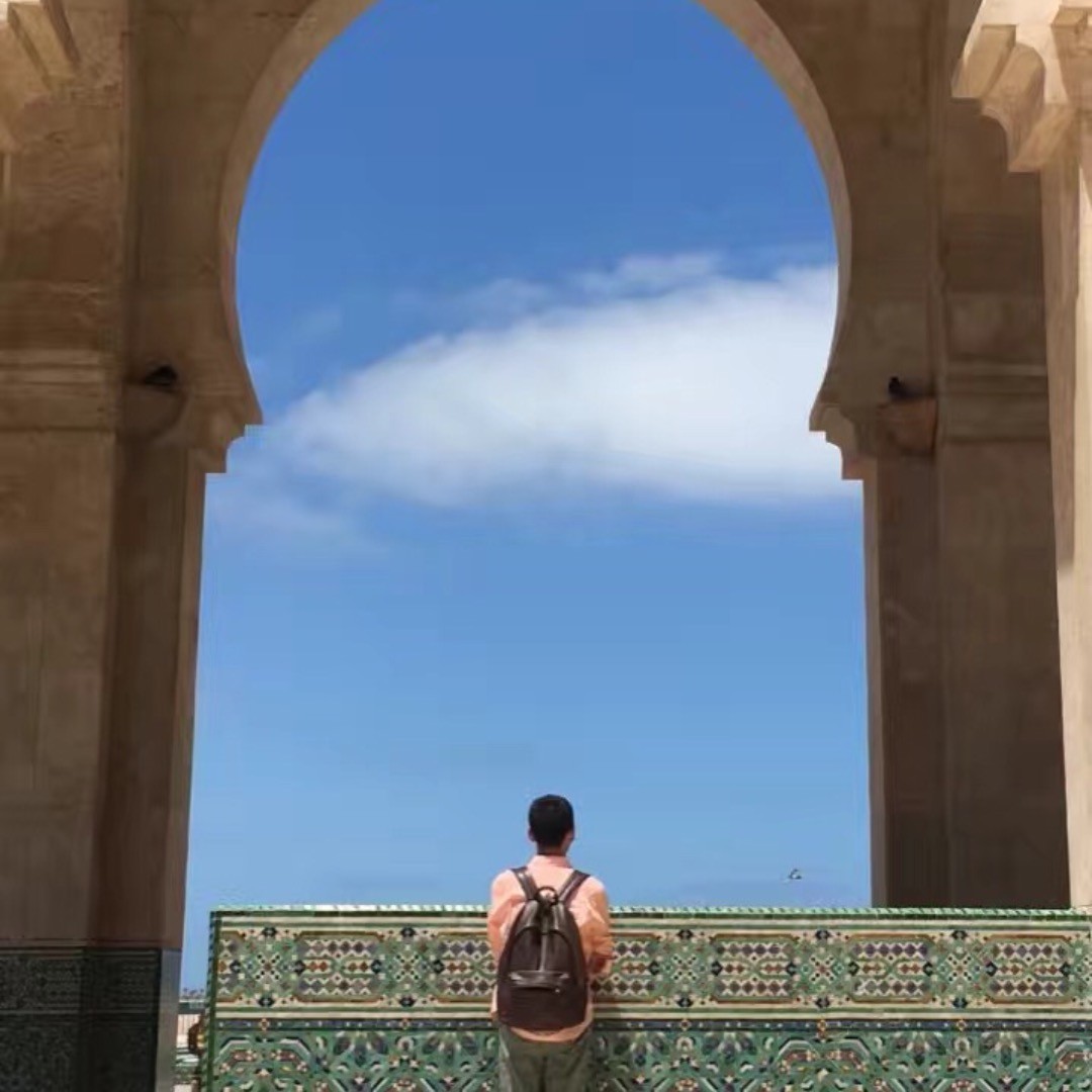 A traveler with a brown leather backpack stands from behind, looking out through a large, towering stone horseshoe arch at the Hassan II Mosque in Casablanca, Morocco. The arch frames a vibrant blue sky with a prominent white cloud, while a decorative tiled balustrade featuring green and gold geometric zellij mosaic patterns runs along the bottom foreground.
